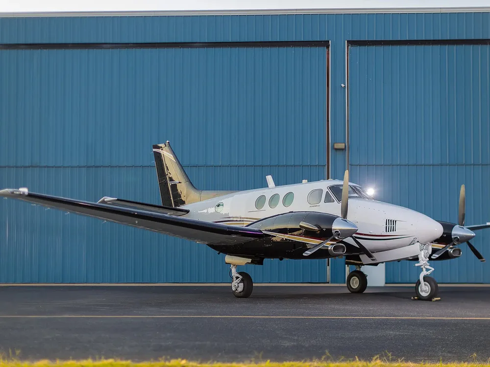 A white and black Beechcraft King Air aircraft with gold and red stripes parked on an tarmac in front of a blue hangar.