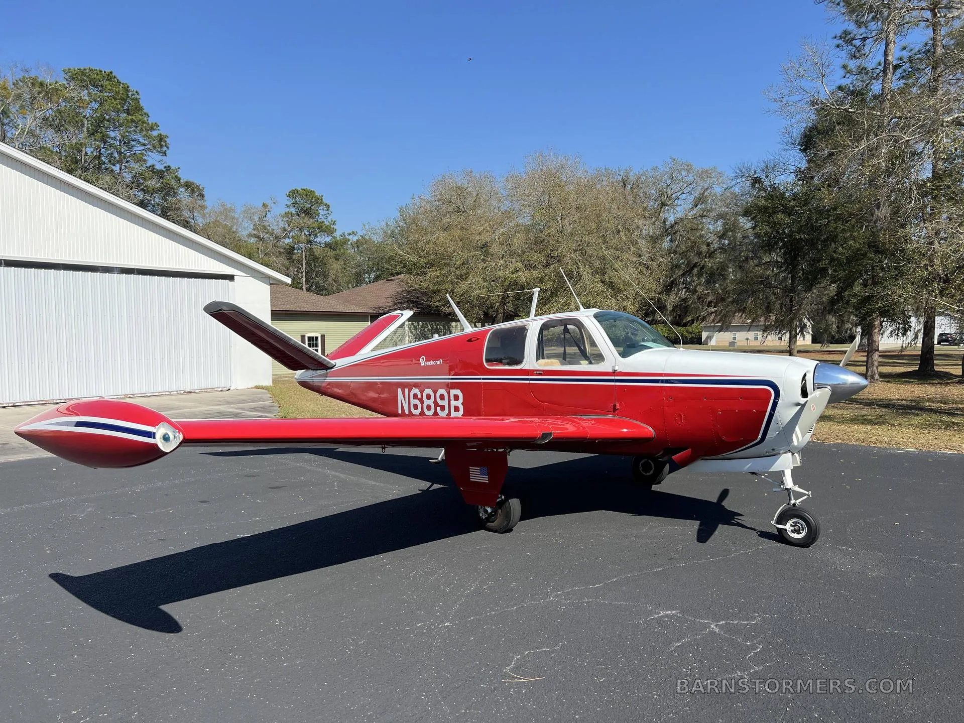 A red and white Beechcraft Bonanza with registration N689B is parked on a driveway next to a white hangar.