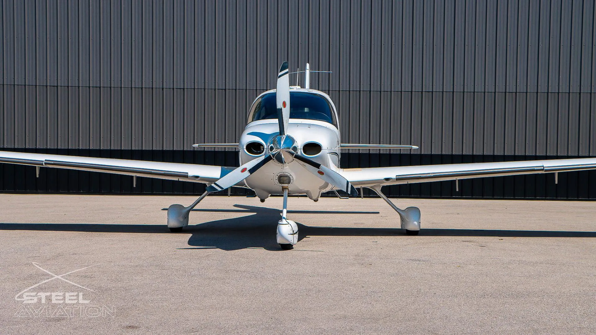 Close-up front view of a white Cirrus SR22 aircraft, showing its propeller and nose, in front of a grey hangar.