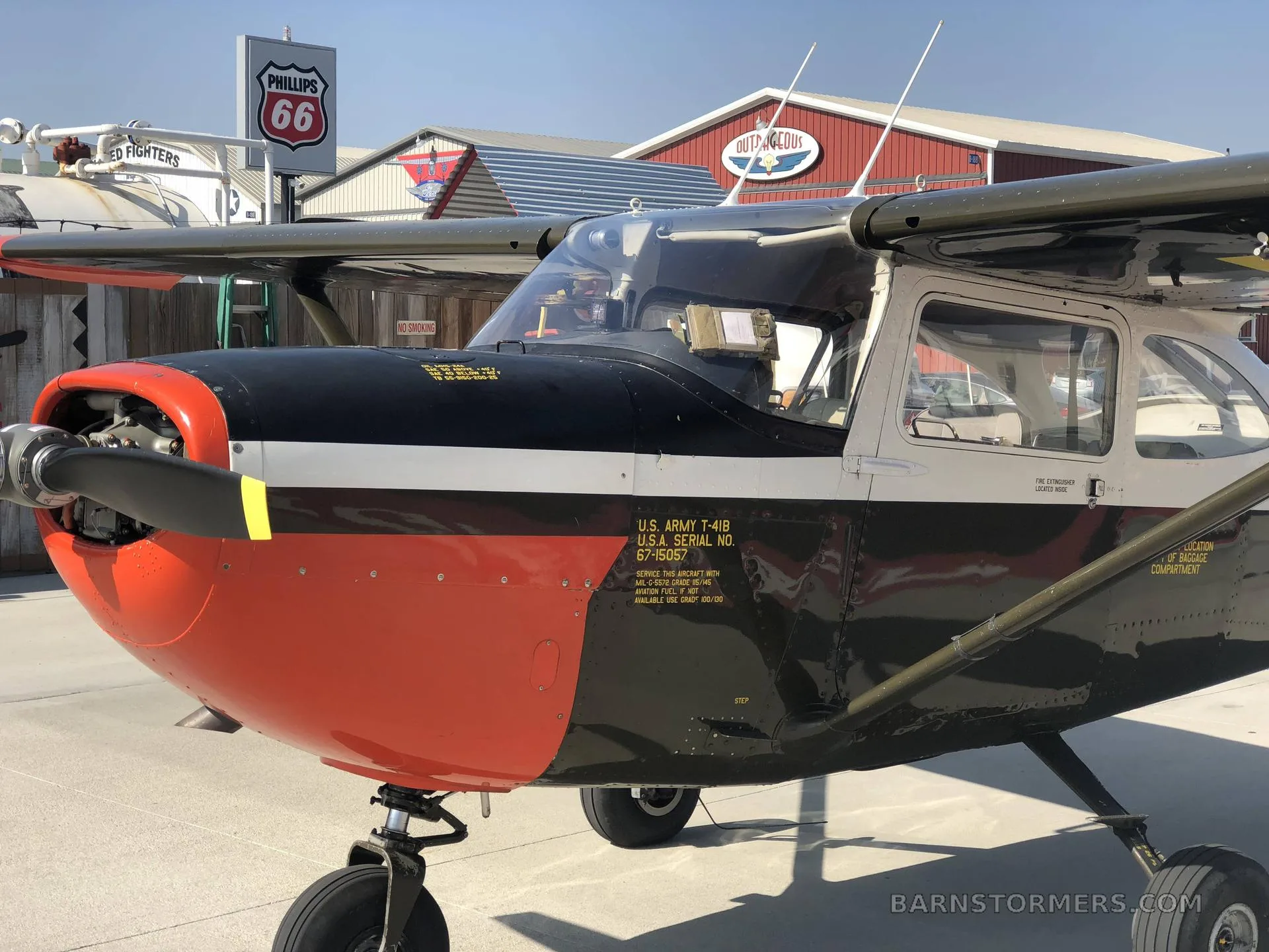 A close-up view of the front and side of a black, white, and red U.S. Army T-41B aircraft parked in front of a building with a Phillips 66 sign.