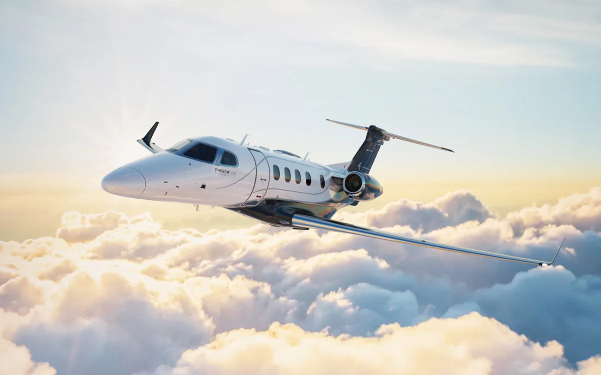 A white Embraer Phenom 300E business jet with dark accents flies above clouds at sunset.