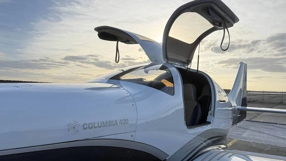A close-up of the cockpit and open gull-wing doors of a white and grey Columbia 400 aircraft at sunset.