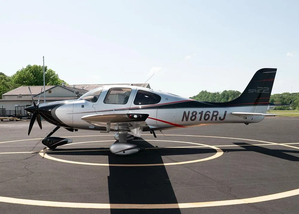 A silver and black Cirrus SR22 aircraft with registration N816RJ is parked on an airfield with trees and buildings in the background.