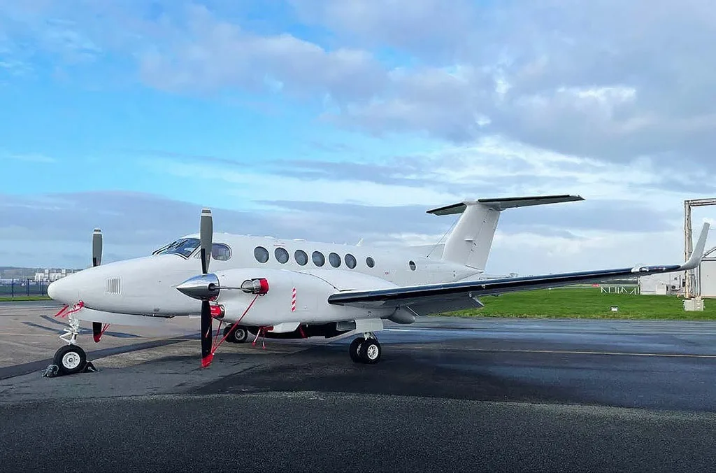 A white twin-engine turboprop aircraft, likely a King Air, parked on an airport tarmac under a partly cloudy sky.