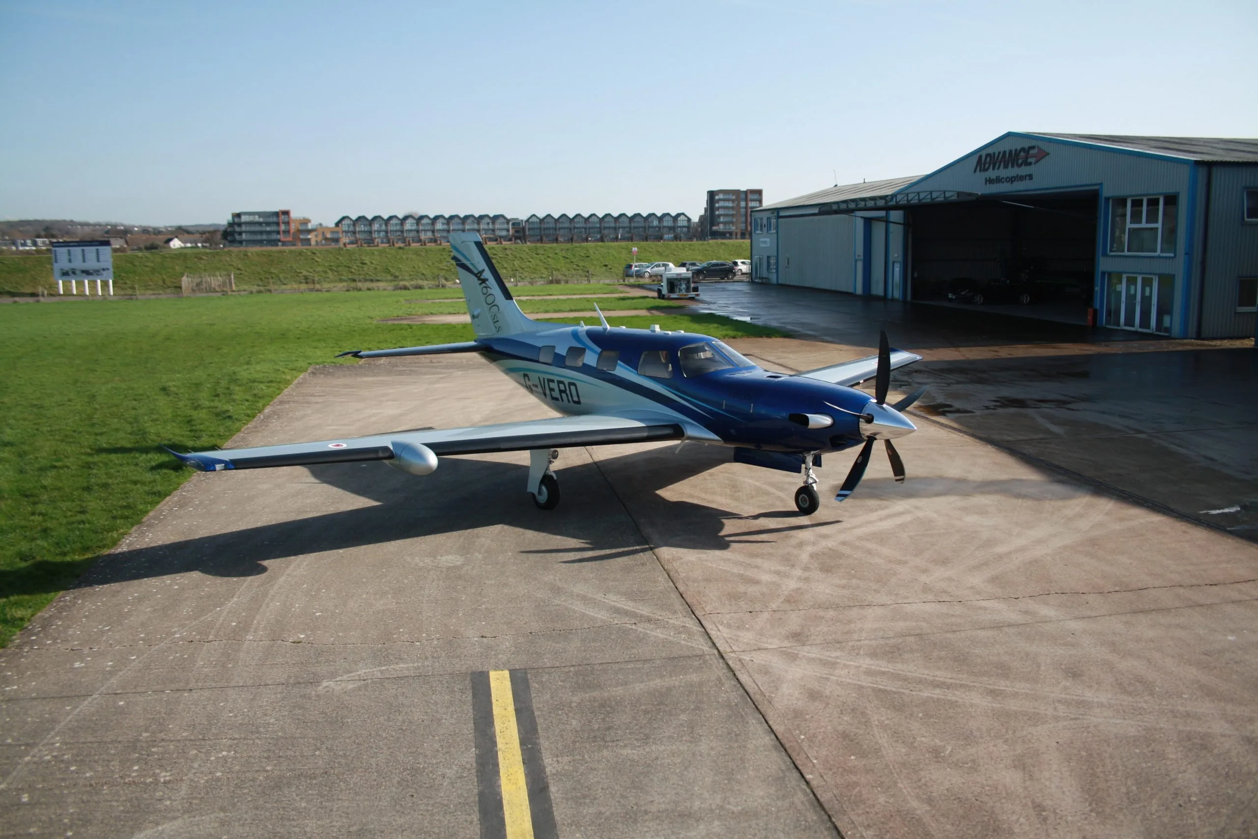 A blue and silver Piper M600SLS aircraft (G-VERO) parked on a tarmac next to an "Advance Helicopters" hangar.