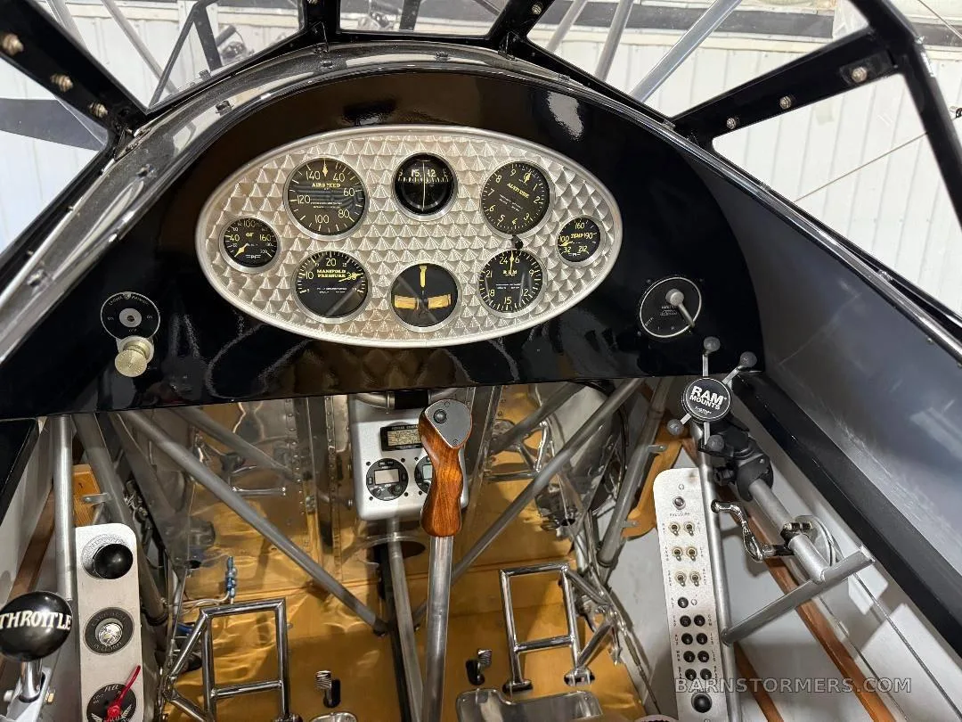 The cockpit of a biplane features a black instrument panel with an oval-shaped cluster of gauges and a wooden control stick.