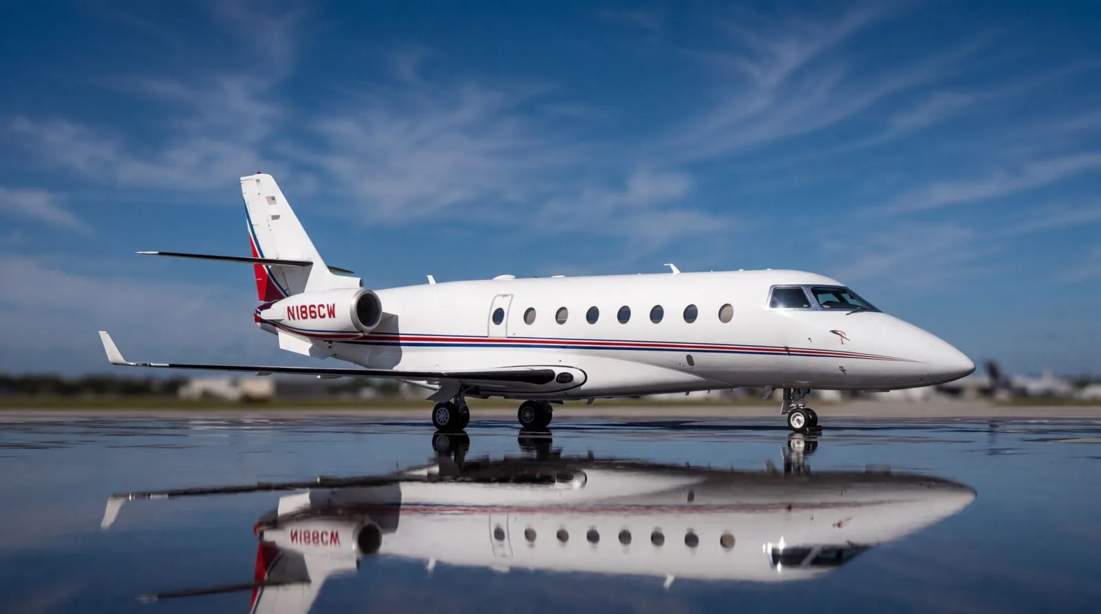 A white business jet with red and blue stripes, registration N186CW, is parked on a wet tarmac, reflecting under a blue sky.