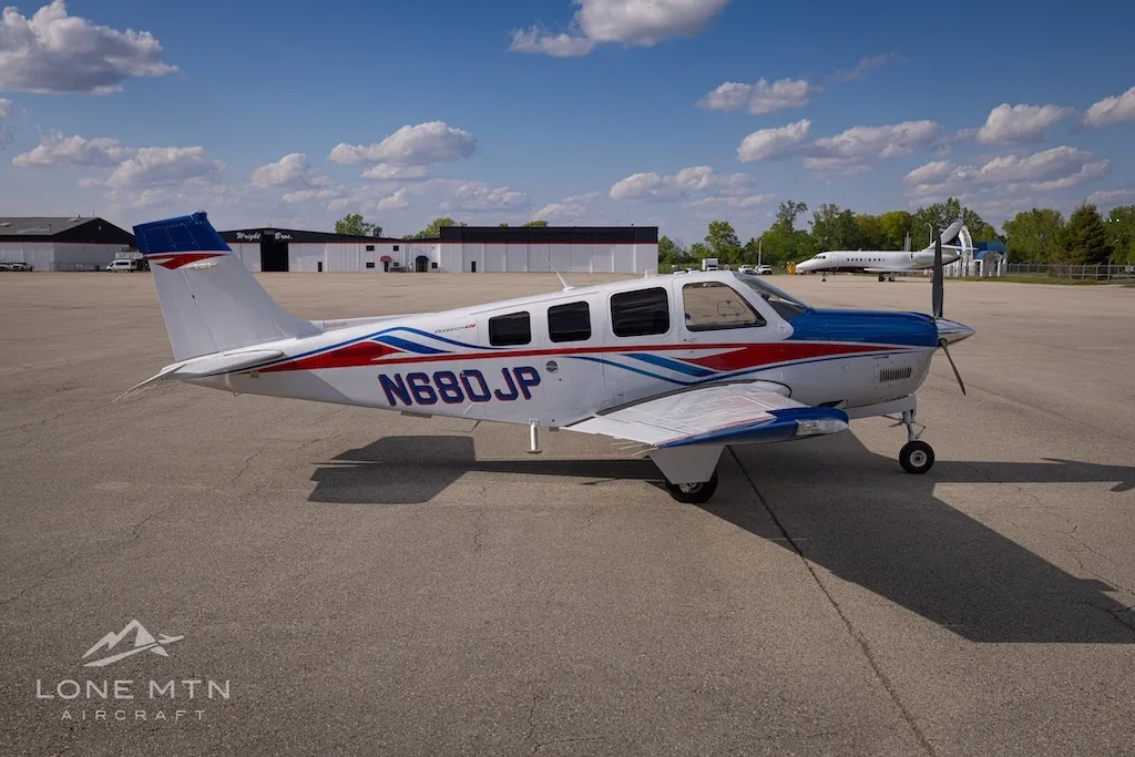 A side view of a white, blue, and red Beechcraft Bonanza N680JP parked on an airfield with other buildings in the background.