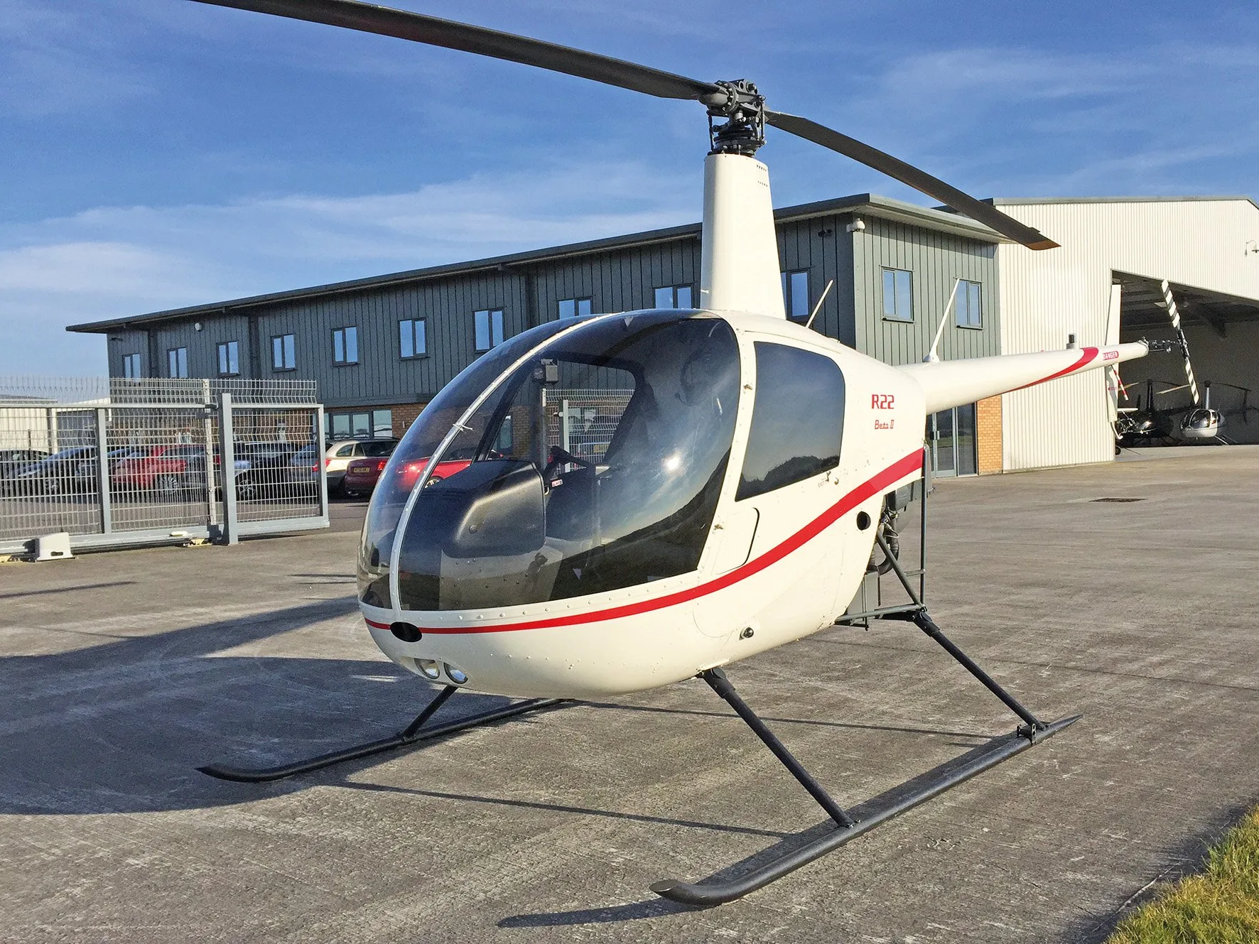 A white Robinson R22 Beta II helicopter with red stripes is parked on an tarmac in front of a hangar.