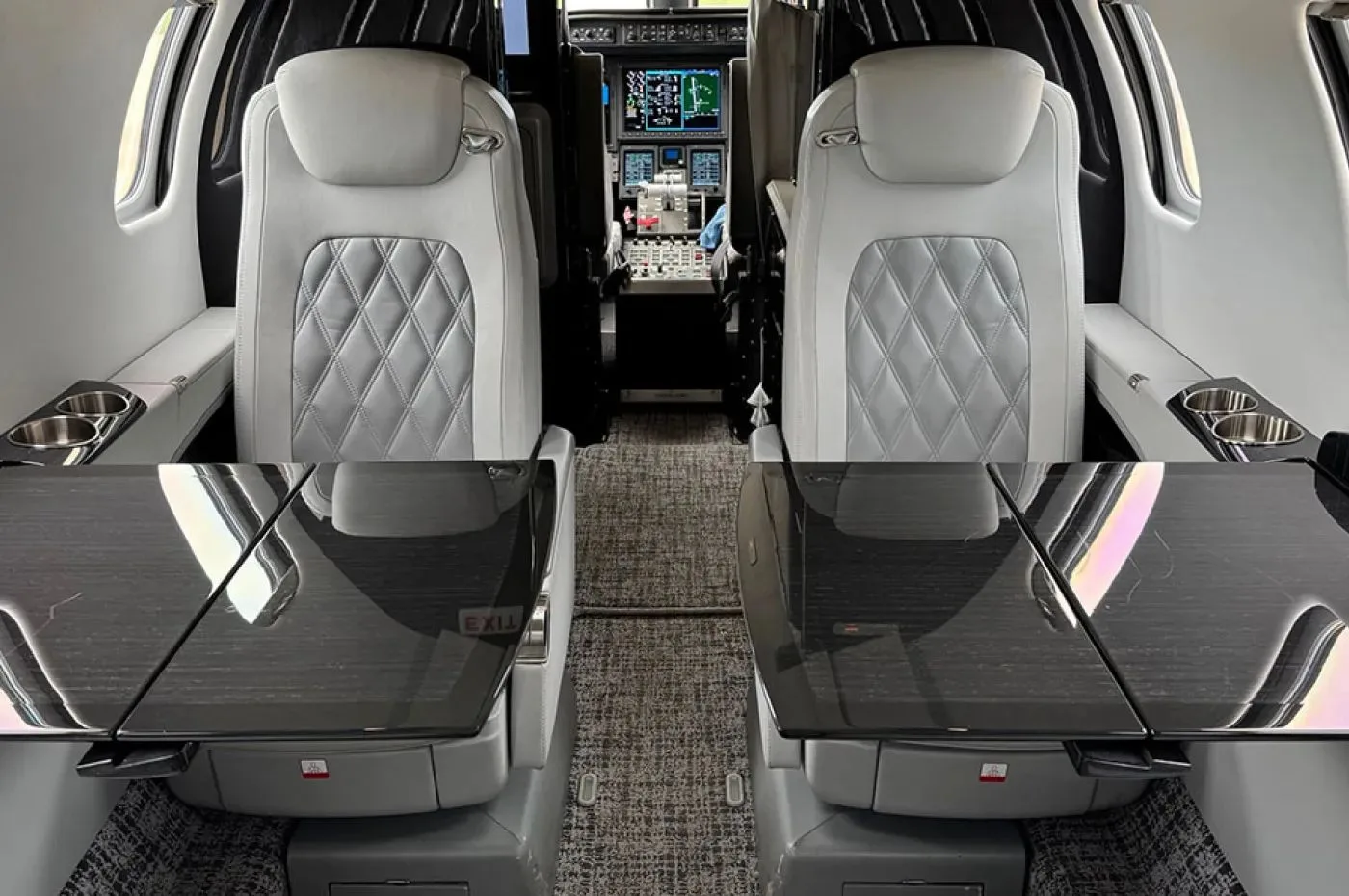 The forward cabin interior of a business jet with light grey quilted leather seats and dark polished tables, looking towards the cockpit.