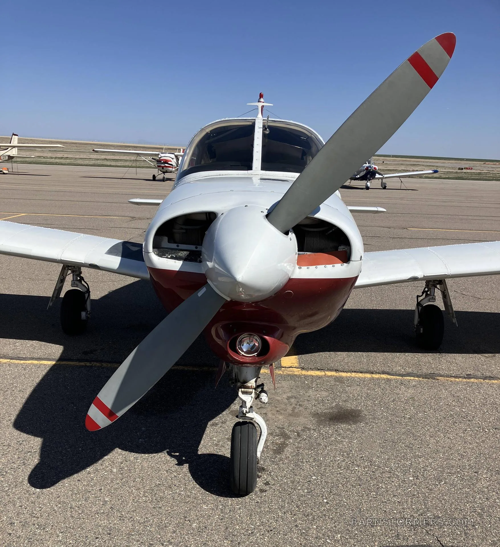 A head-on view of the white and maroon Piper Arrow aircraft with its propeller, parked on an airport tarmac.