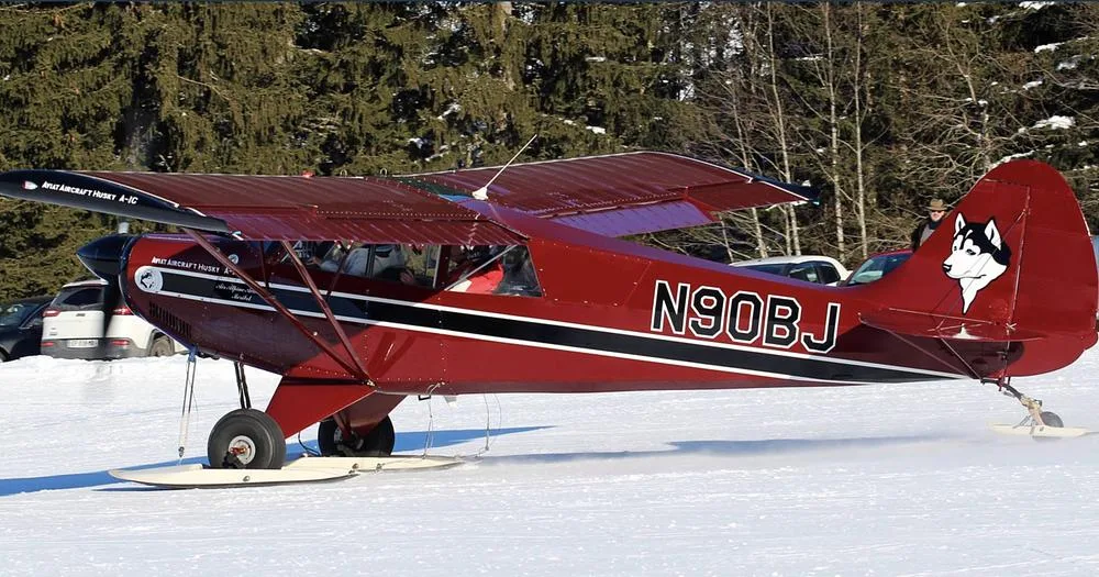 A burgundy Aviat Aircraft Husky A-1C, registration N90BJ, equipped with skis, on a snowy field with trees in the background.
