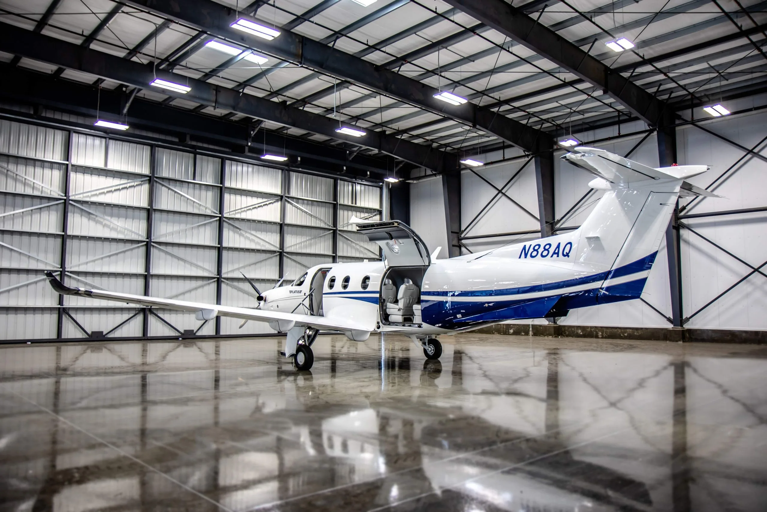 A white Pilatus PC-12 with blue stripes and open passenger doors is parked inside a hangar with a reflective floor.