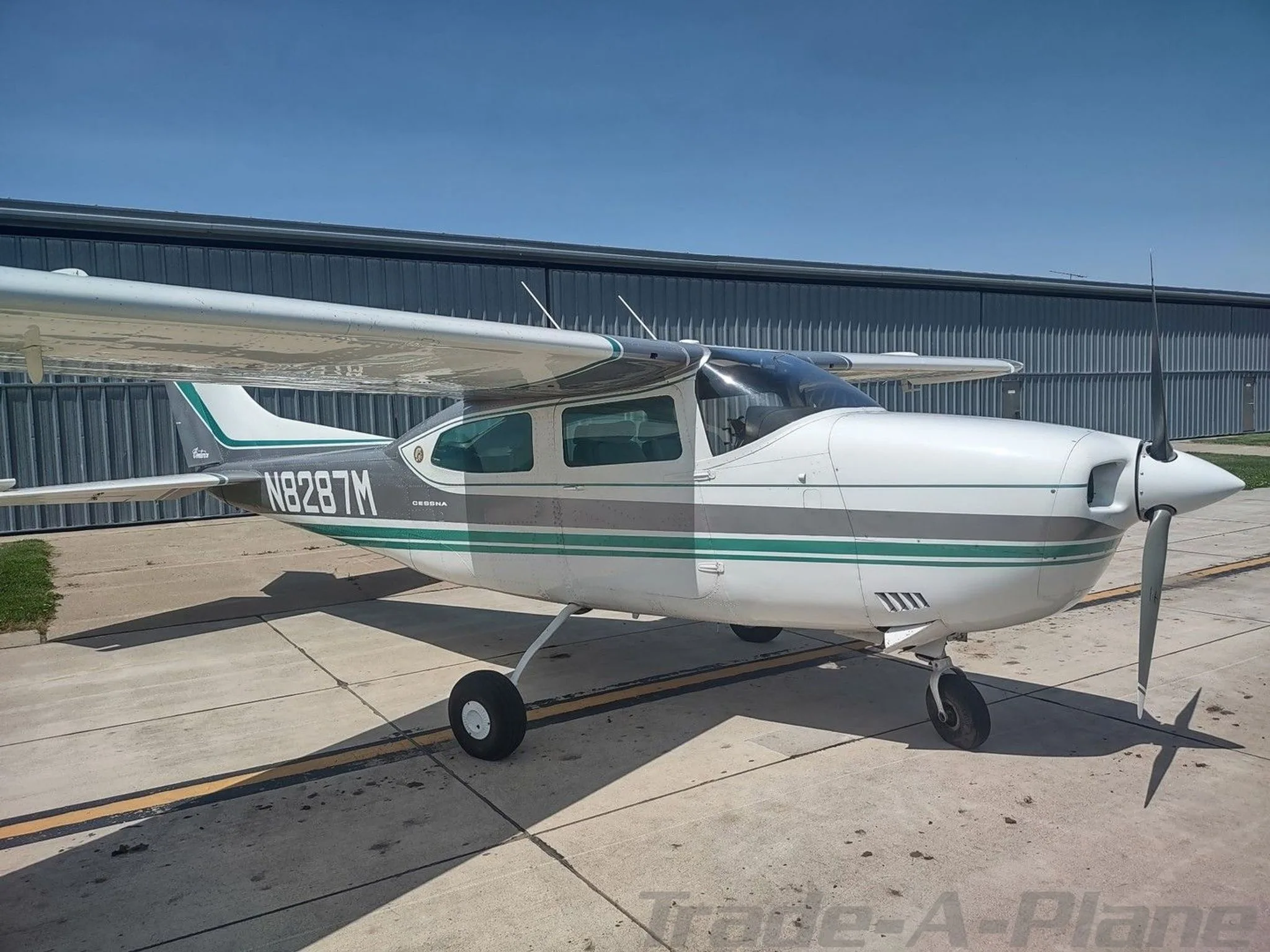 A white and grey Cessna aircraft with green stripes, registration N8287M, parked on a tarmac with its propeller visible.