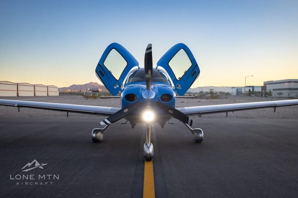 A head-on view of a blue and silver Cirrus SR22 aircraft with open doors and landing light on, parked on an airfield.