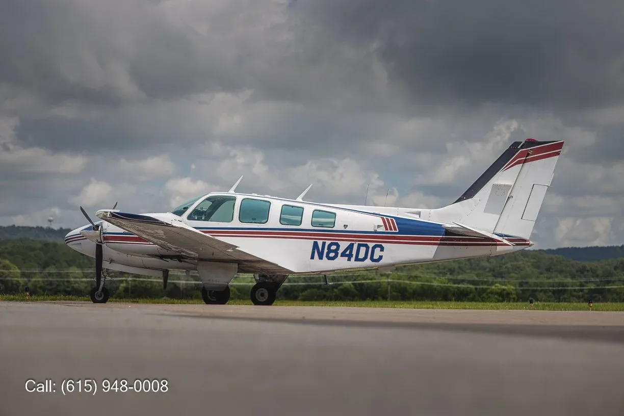 A white twin-engine propeller aircraft with blue and red stripes, registration N84DC, parked on an airfield under a cloudy sky.