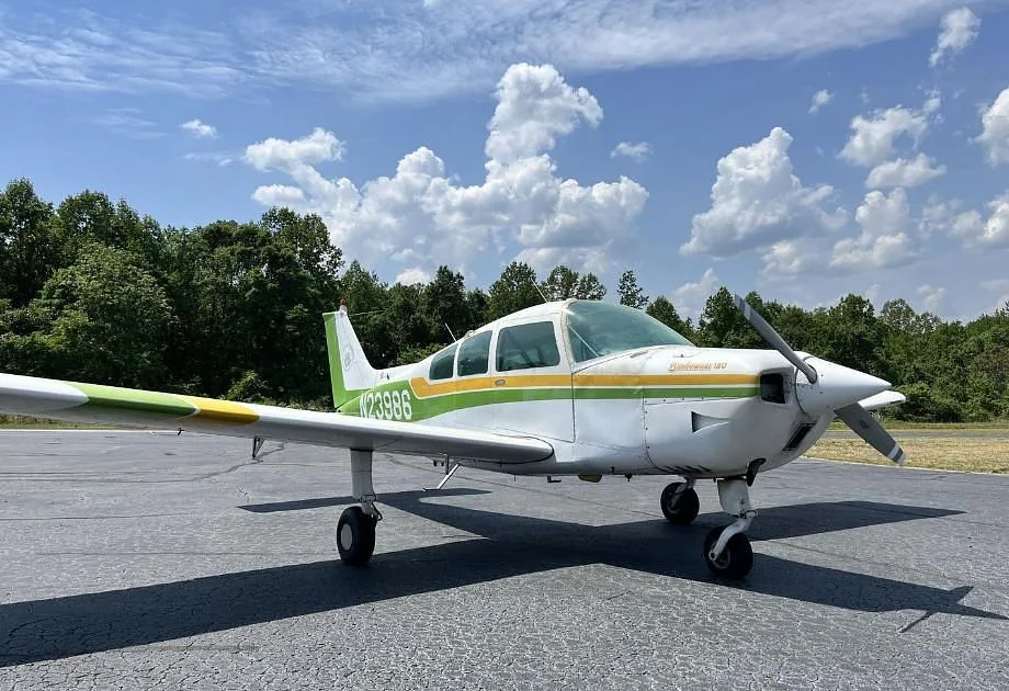A white and green Beechcraft Sundowner 180 (N23986) parked on an asphalt runway under a partly cloudy sky.