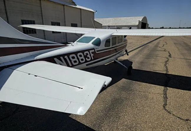 An overhead view of the tail and right wing of a white and maroon Cessna 210 Centurion aircraft with registration N1883F, parked near hangars.