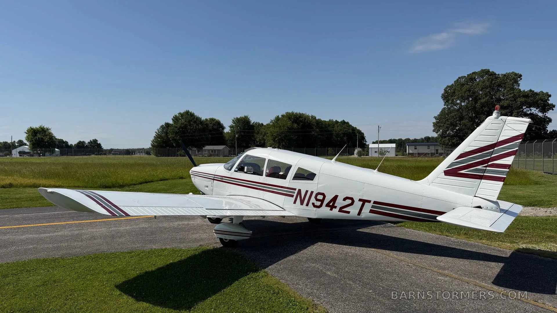 A white single-engine propeller aircraft (N1942T) with red and grey stripes, viewed from the side, parked on an asphalt taxiway next to a grassy field.