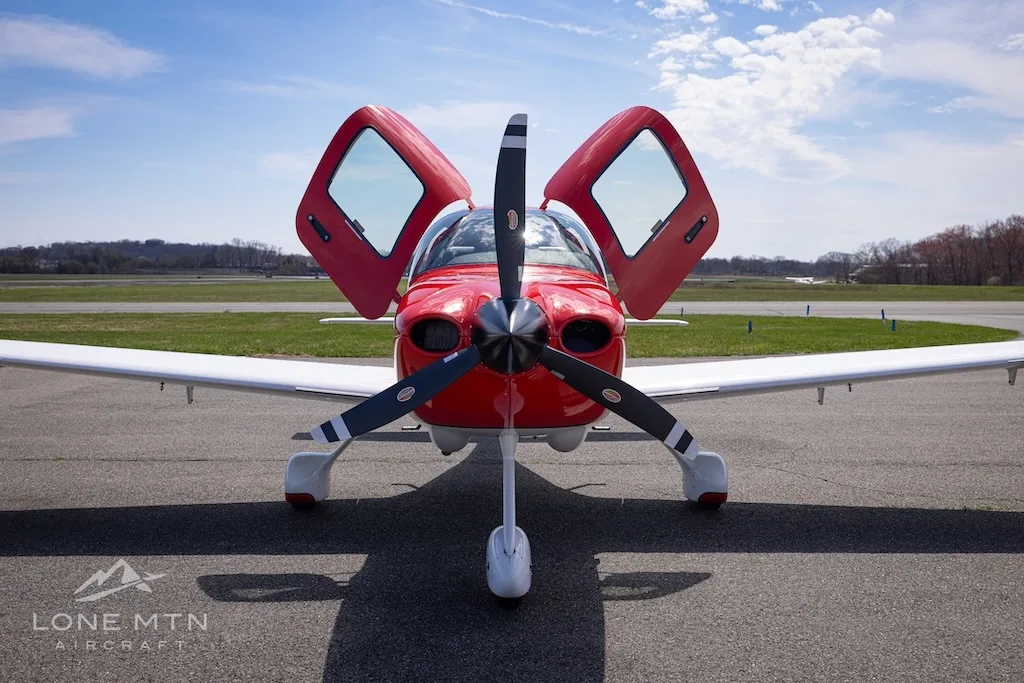A front view of a red and white Cirrus SR20 aircraft with its doors open, parked on an airfield.