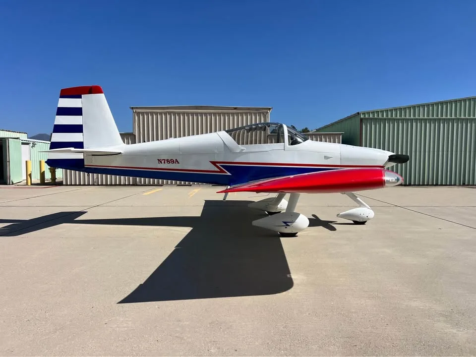 A white, blue, and red single-engine aircraft with a red propeller spinner and striped tail, parked on an airfield.