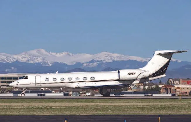 A white Gulfstream G550 business jet with black and gold accents on the tail, taxiing on a runway with snow-capped mountains in the background.