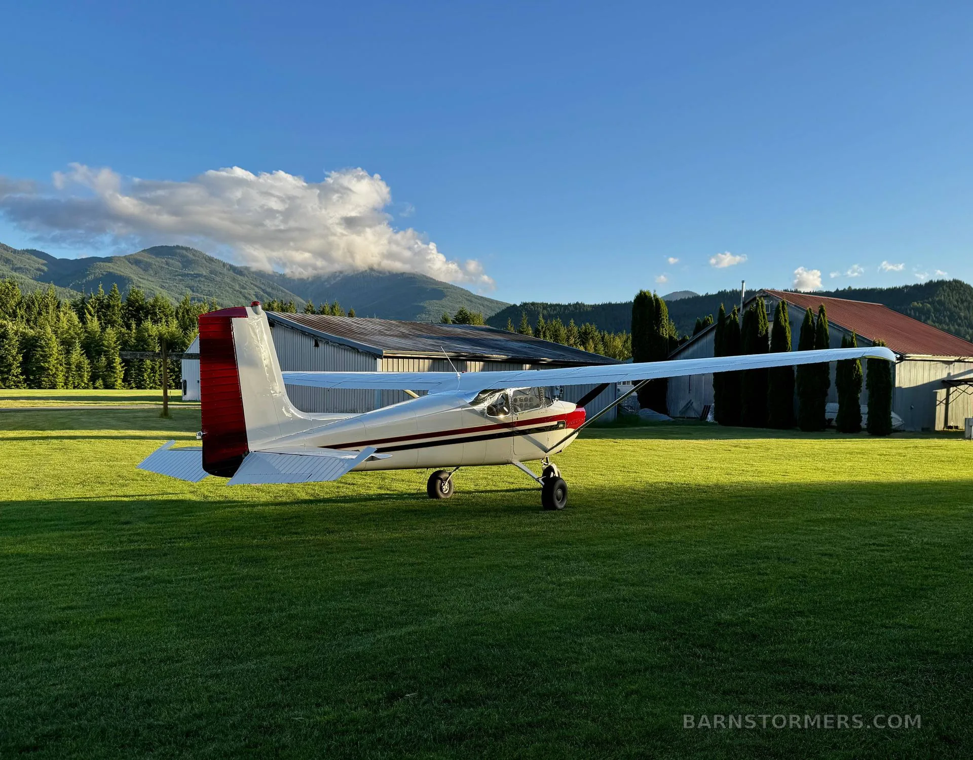 A white and red Cessna 172 aircraft is parked on a green grassy field, viewed from the rear-left, with hangars and mountains in the background.