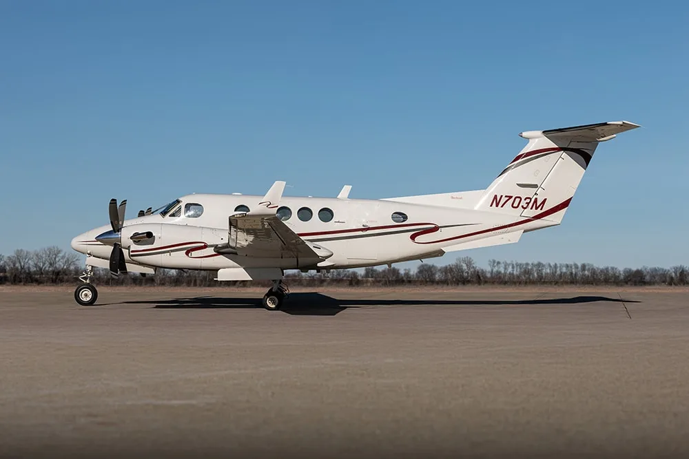 A white twin-turboprop aircraft with maroon and black stripes, registration N703M, parked on an airfield under a clear blue sky.