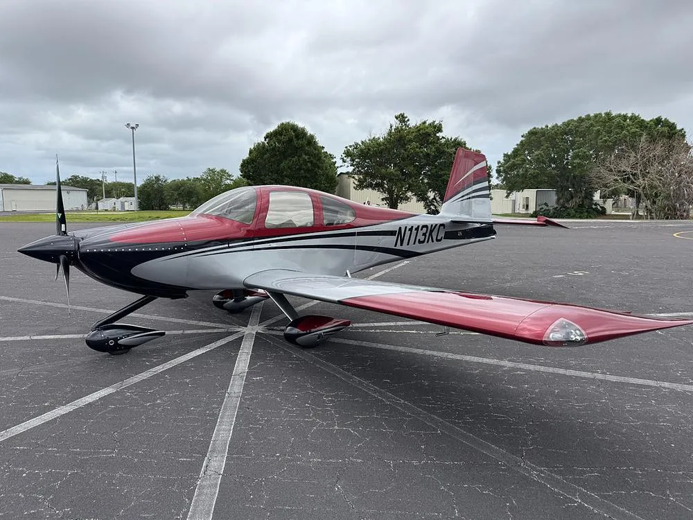 A red, silver, and black single-engine aircraft (N113KC) parked on an asphalt tarmac under a cloudy sky.