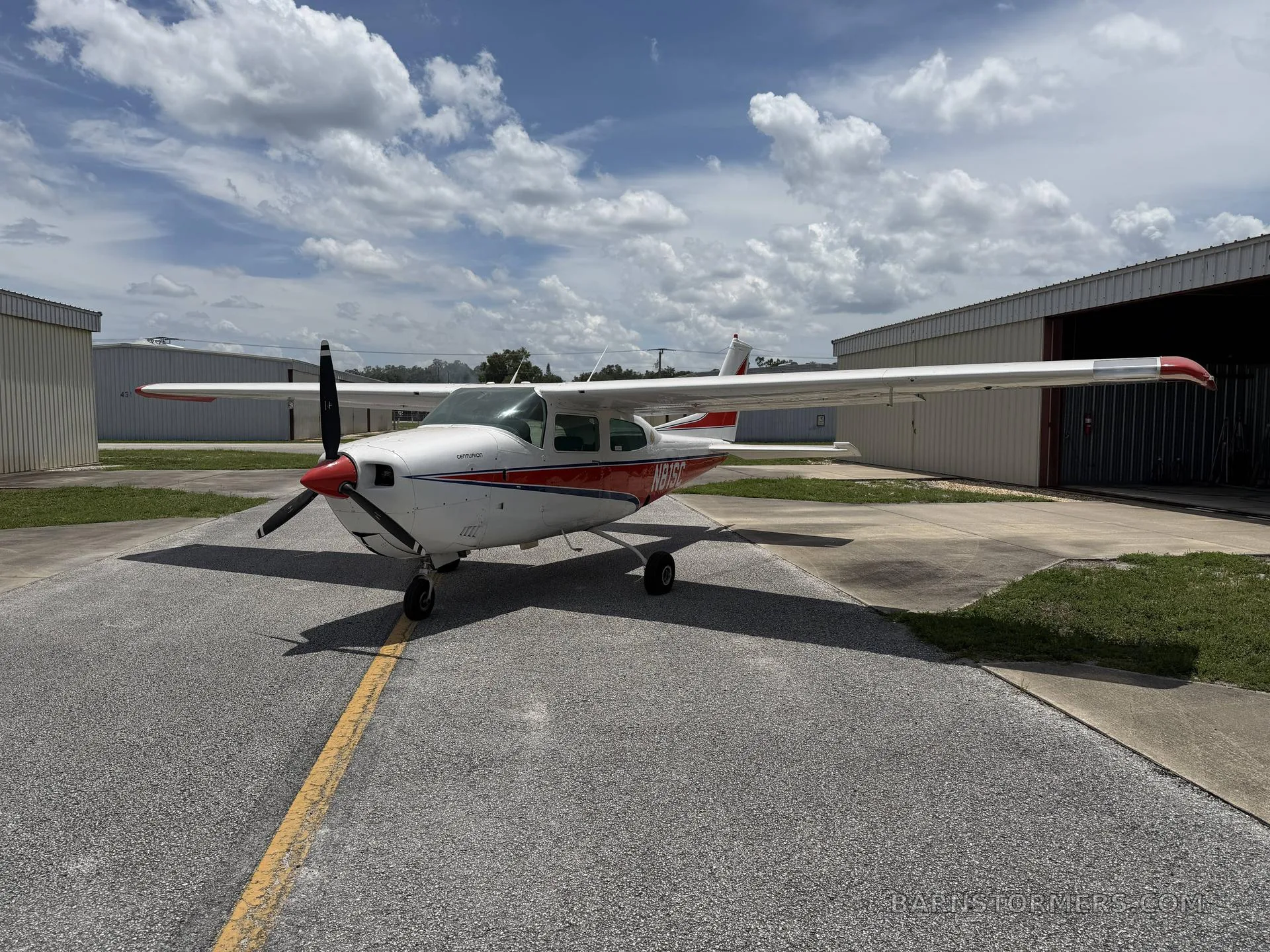 A white and red Cessna 210 Centurion aircraft is parked on an asphalt ramp between hangars under a partly cloudy sky.