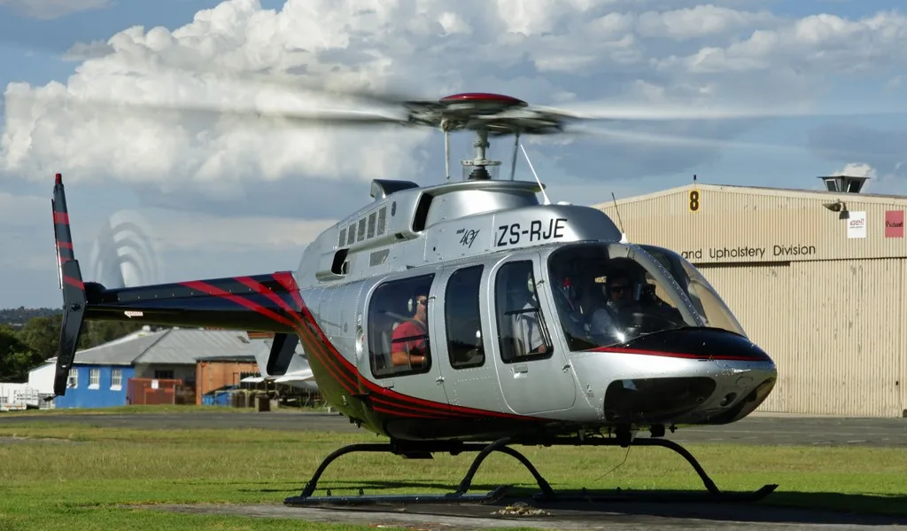 A silver Bell 407 helicopter with black and red stripes sits on a grassy airfield with a hangar in the background.