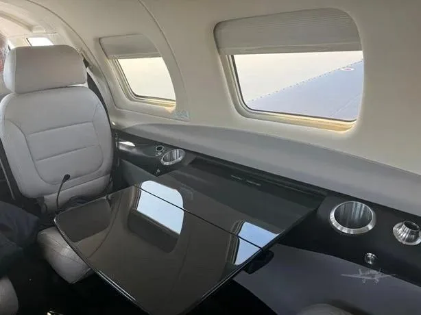 The interior cabin of an aircraft with light-colored seats, windows, and a dark, reflective fold-out table.