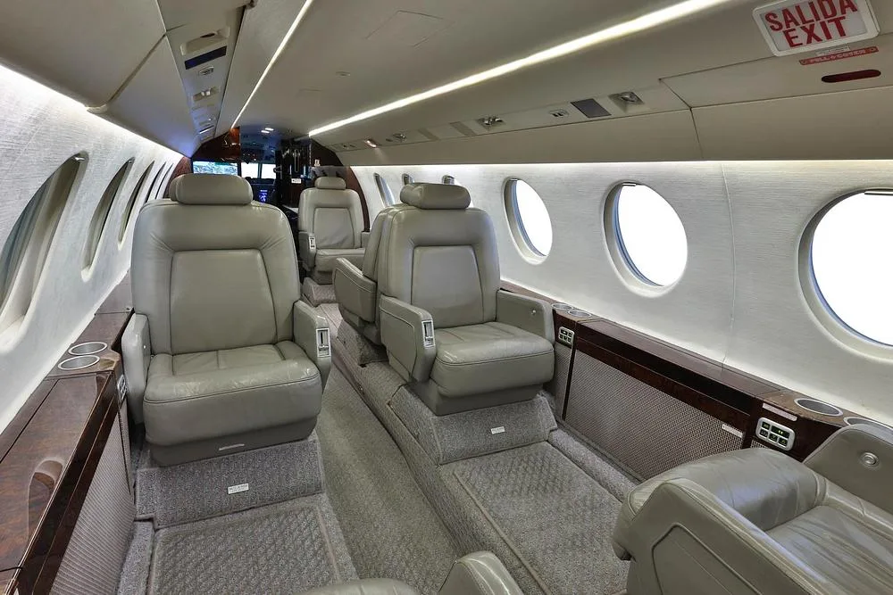 A view of the business jet cabin interior, featuring grey leather seats and large windows.
