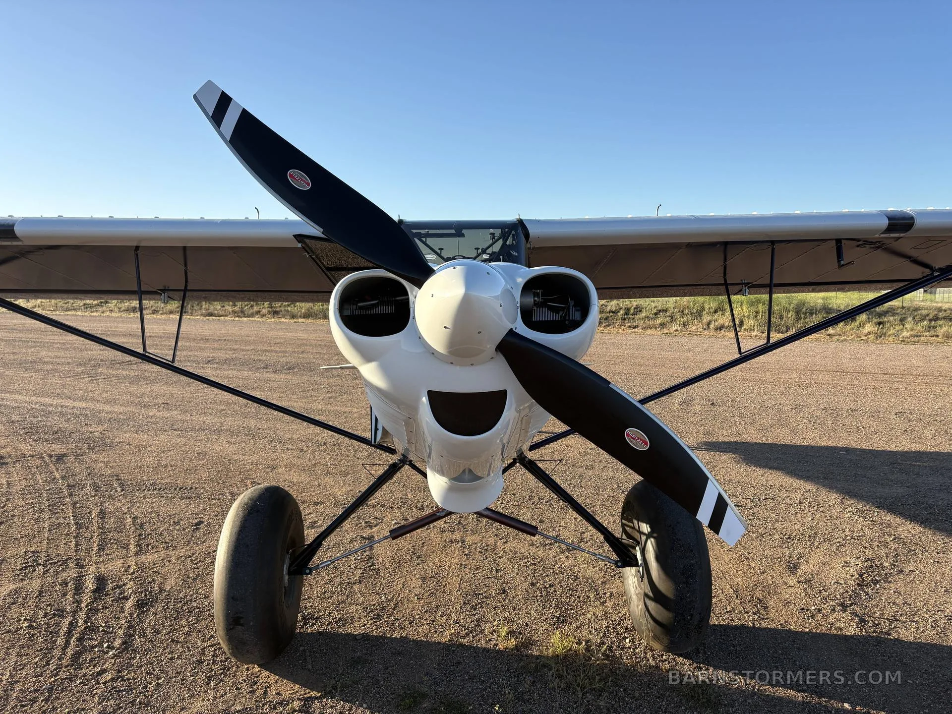 A head-on view of a white and blue CubCrafters aircraft with a large propeller and tundra tires on a dirt field.
