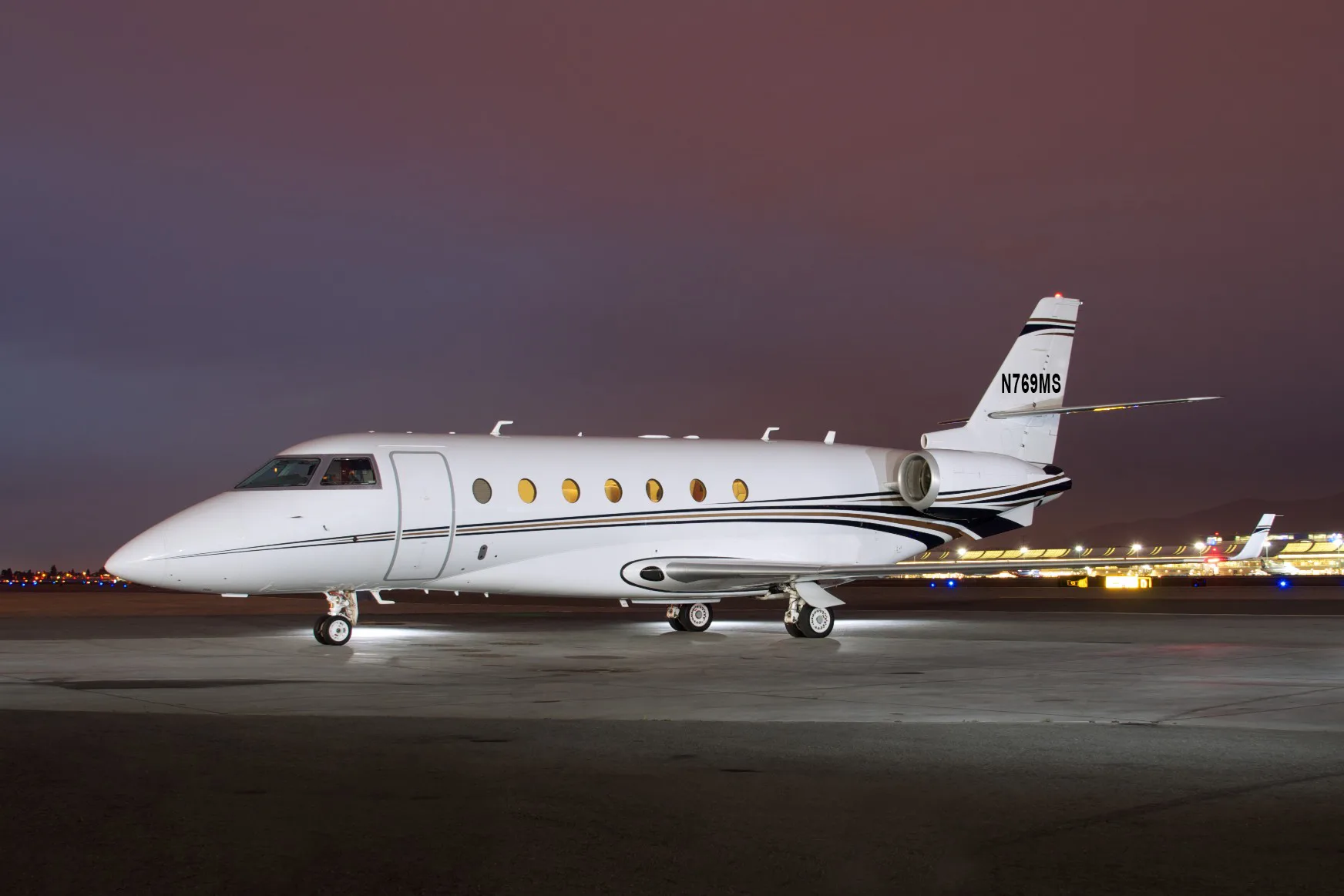 A white business jet with black and gold stripes on the fuselage and tail, parked on a tarmac at night with airport lights in the background.