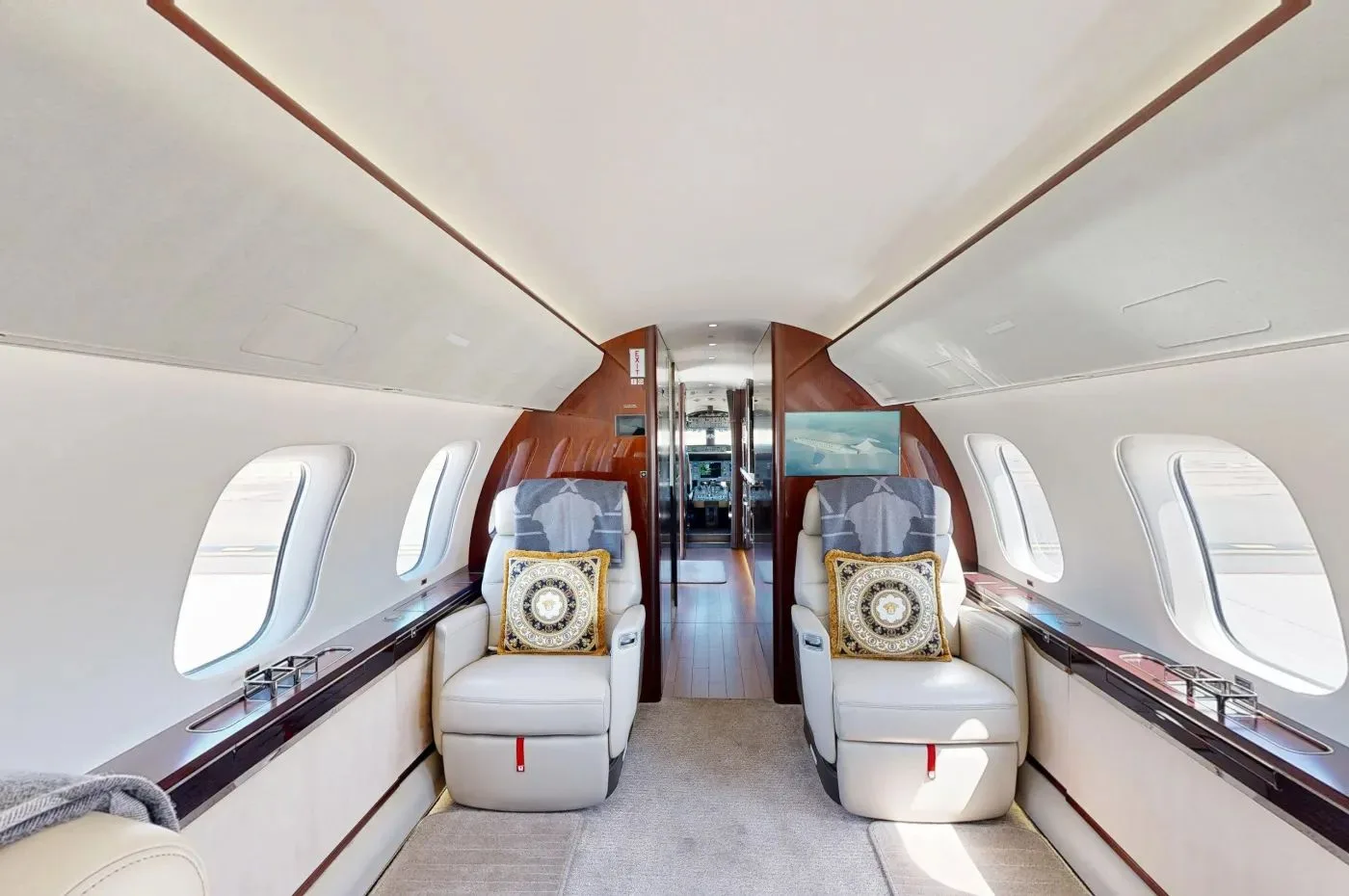 The luxurious cabin interior of a private jet, featuring two cream leather seats with decorative pillows and dark wood accents.