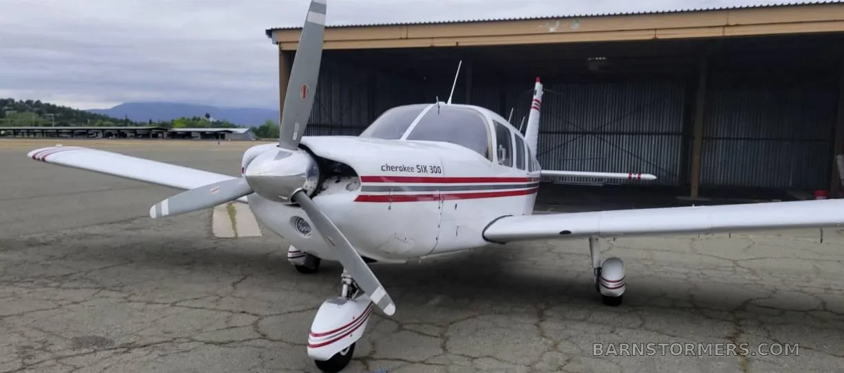 A white Piper Cherokee Six 300 aircraft with red and grey stripes, seen from the front-left, parked on an asphalt apron in front of a hangar.