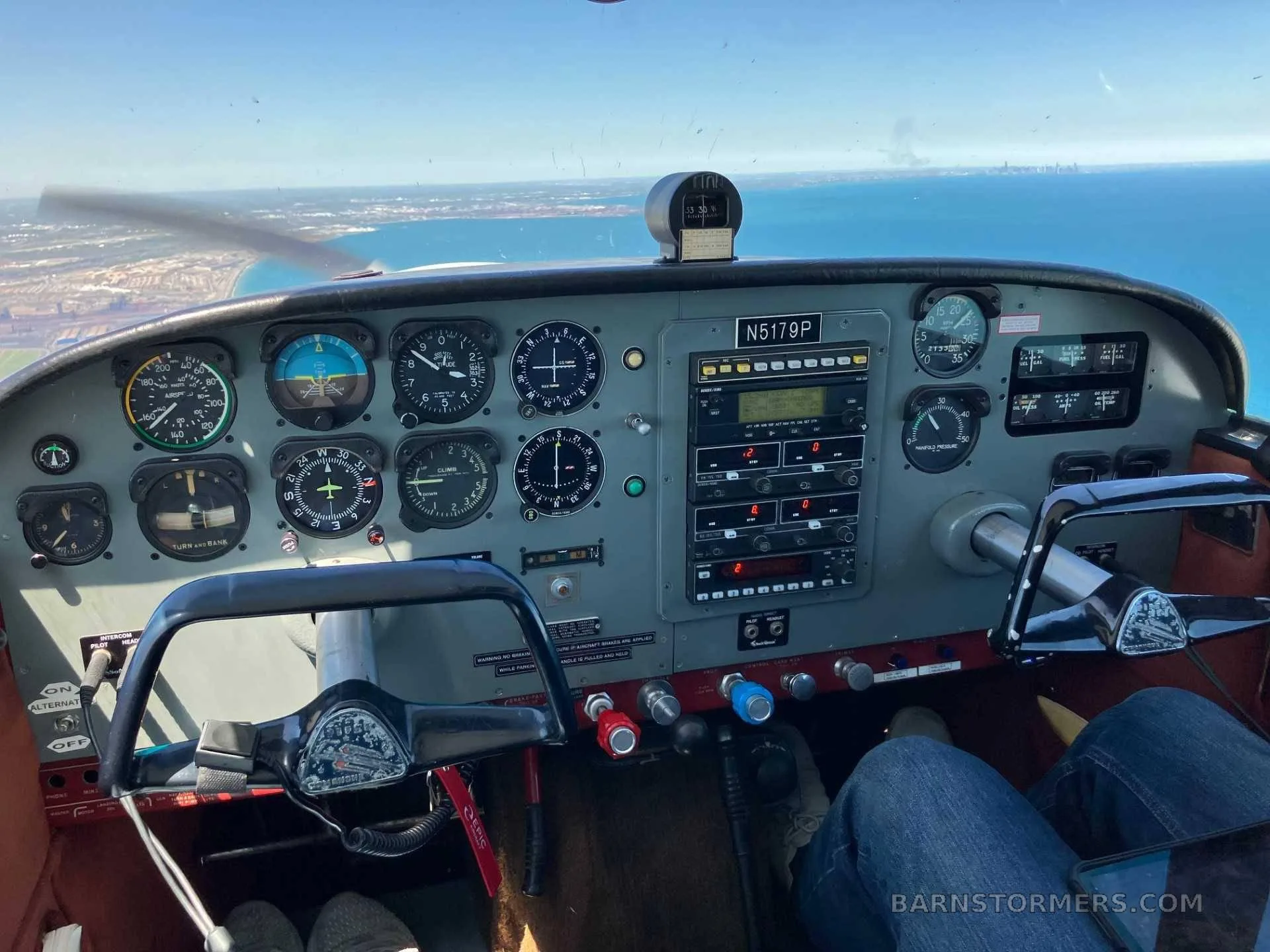 The cockpit of a single-engine aircraft, showing the instrument panel, controls, and a view of a city along a coastline from above.