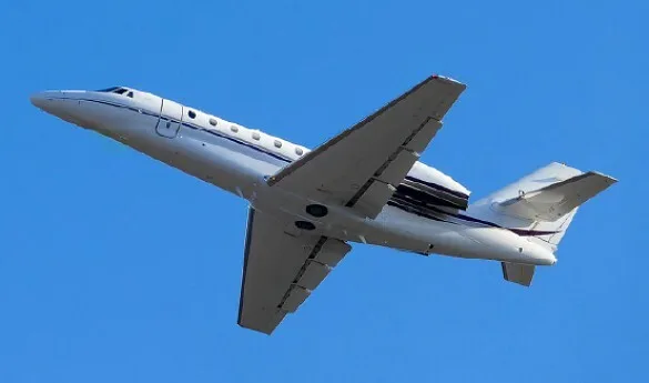 A white business jet with a dark stripe on its fuselage is shown in flight against a clear blue sky.