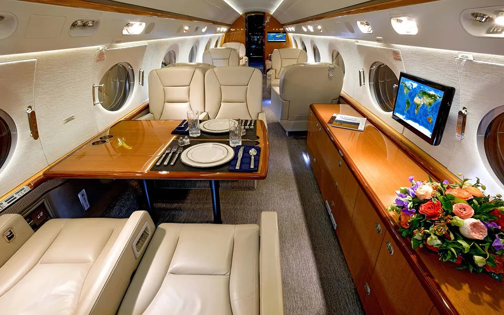 A wide view of the aircraft's luxurious cabin featuring a dining table set for two, a long wooden cabinet, and a bouquet of colorful flowers.