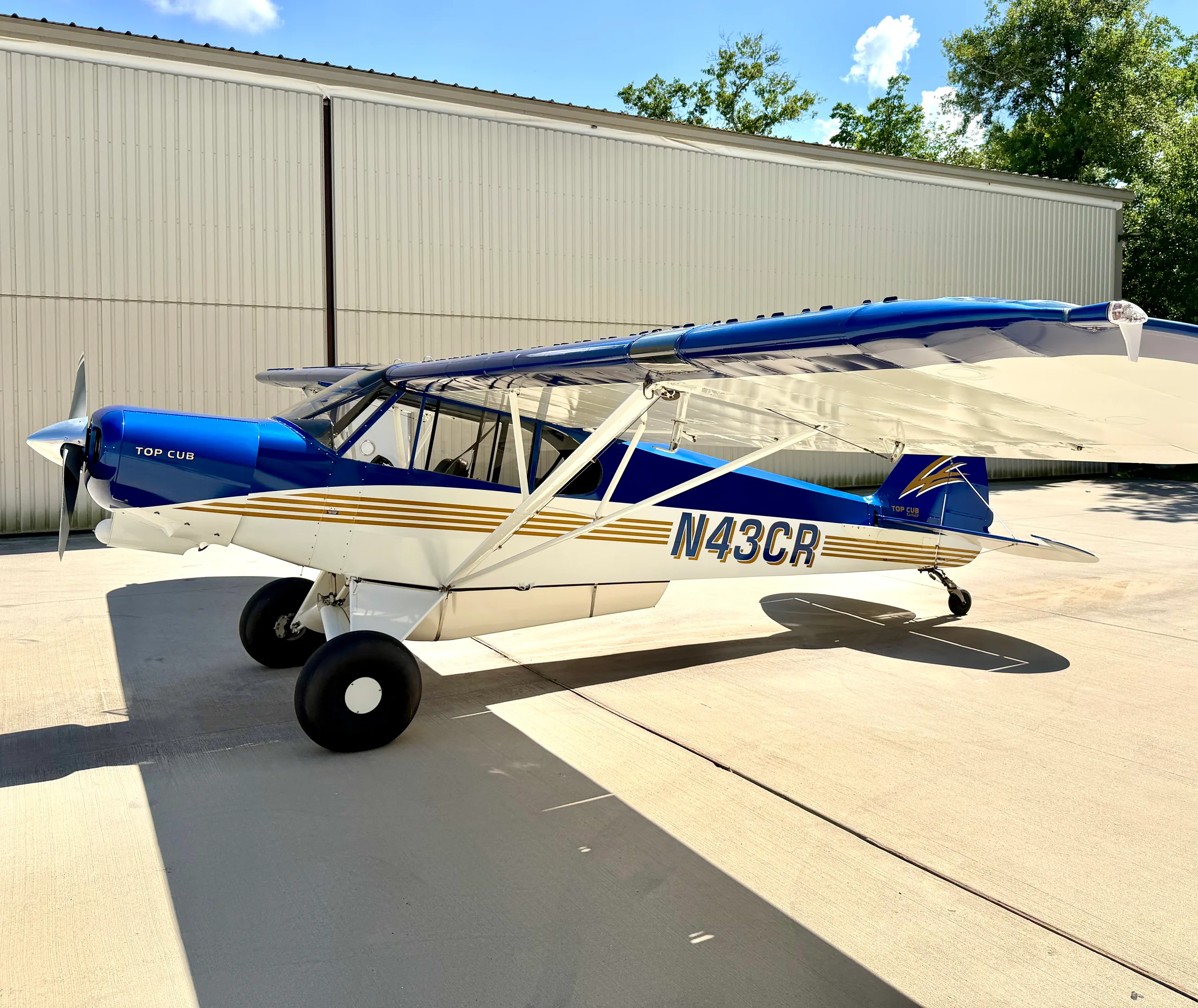 A blue and white Top Cub aircraft with gold stripes is parked on a tarmac in front of a white hangar under a blue sky.