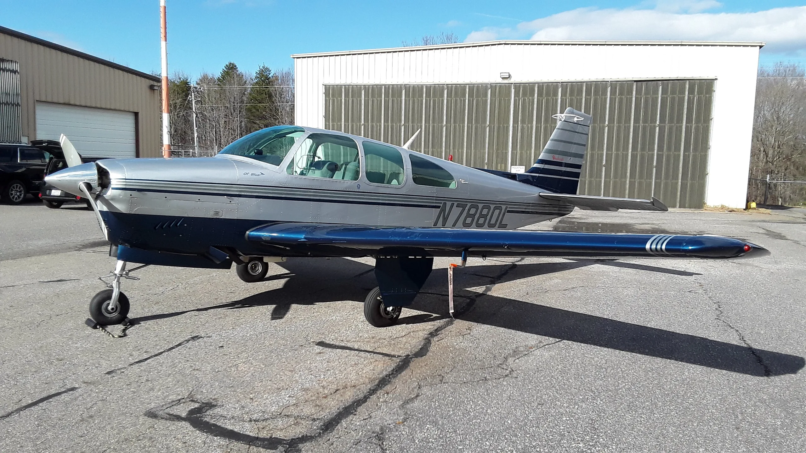 A silver and blue Grumman American AA-5 Traveler aircraft, tail number N7880L, parked on an asphalt apron next to hangars under a clear sky.