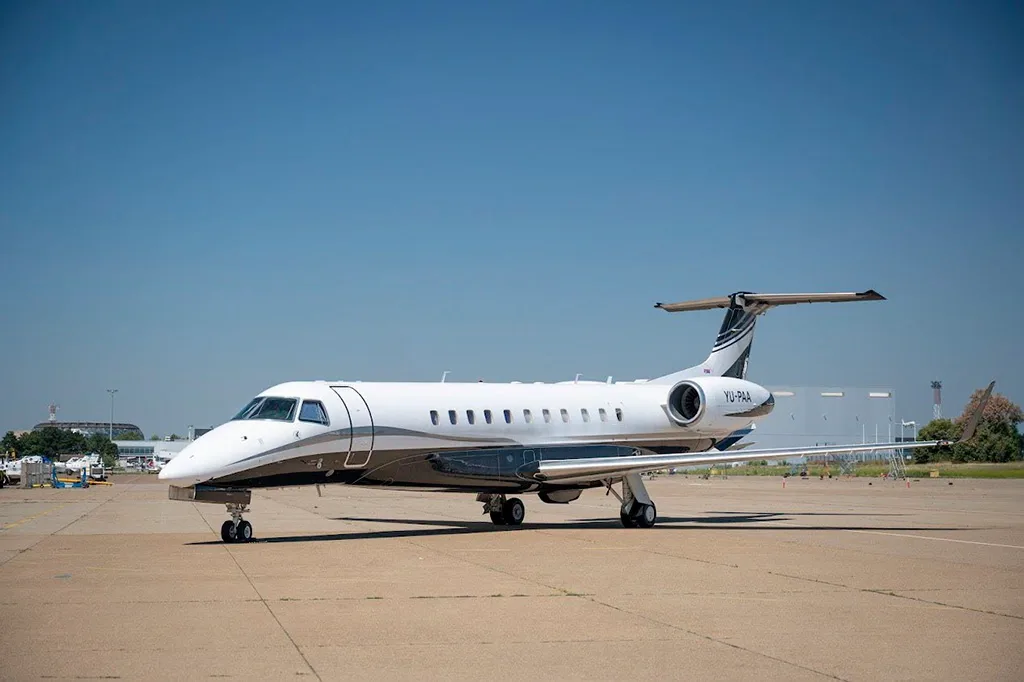 A white and black Embraer Legacy 600 business jet with registration YU-PAA parked on a tarmac under a clear blue sky.