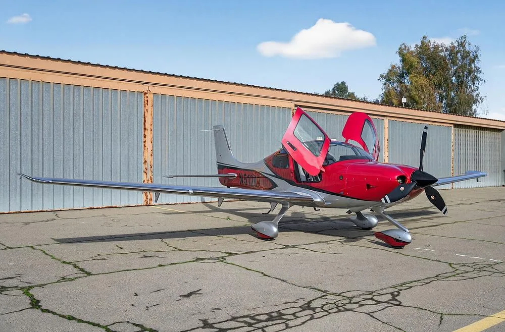 A red and silver single-engine aircraft, N512KP, with its gull-wing doors open, parked on an airfield.