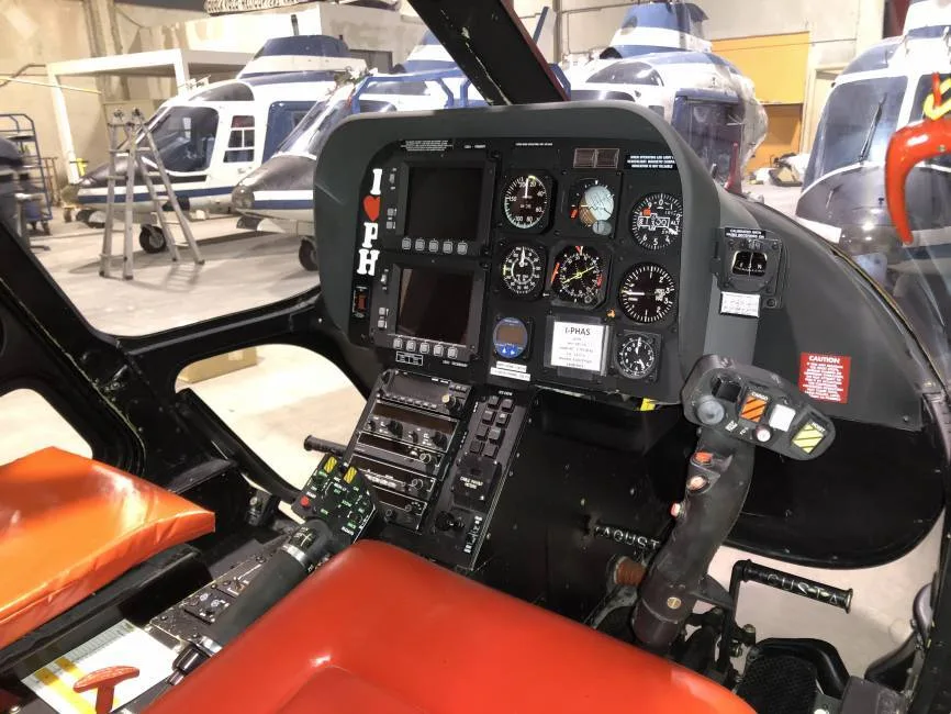 The cockpit of a helicopter with a full instrument panel and controls, with other helicopters visible in the background.