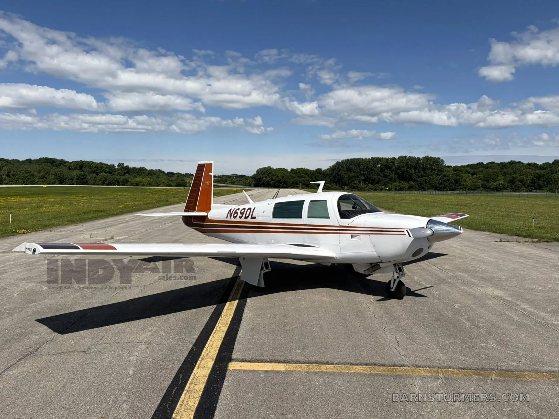 A white and brown Mooney M20J aircraft (N69DL) is parked on an asphalt runway under a partly cloudy sky, viewed from the front-right side.