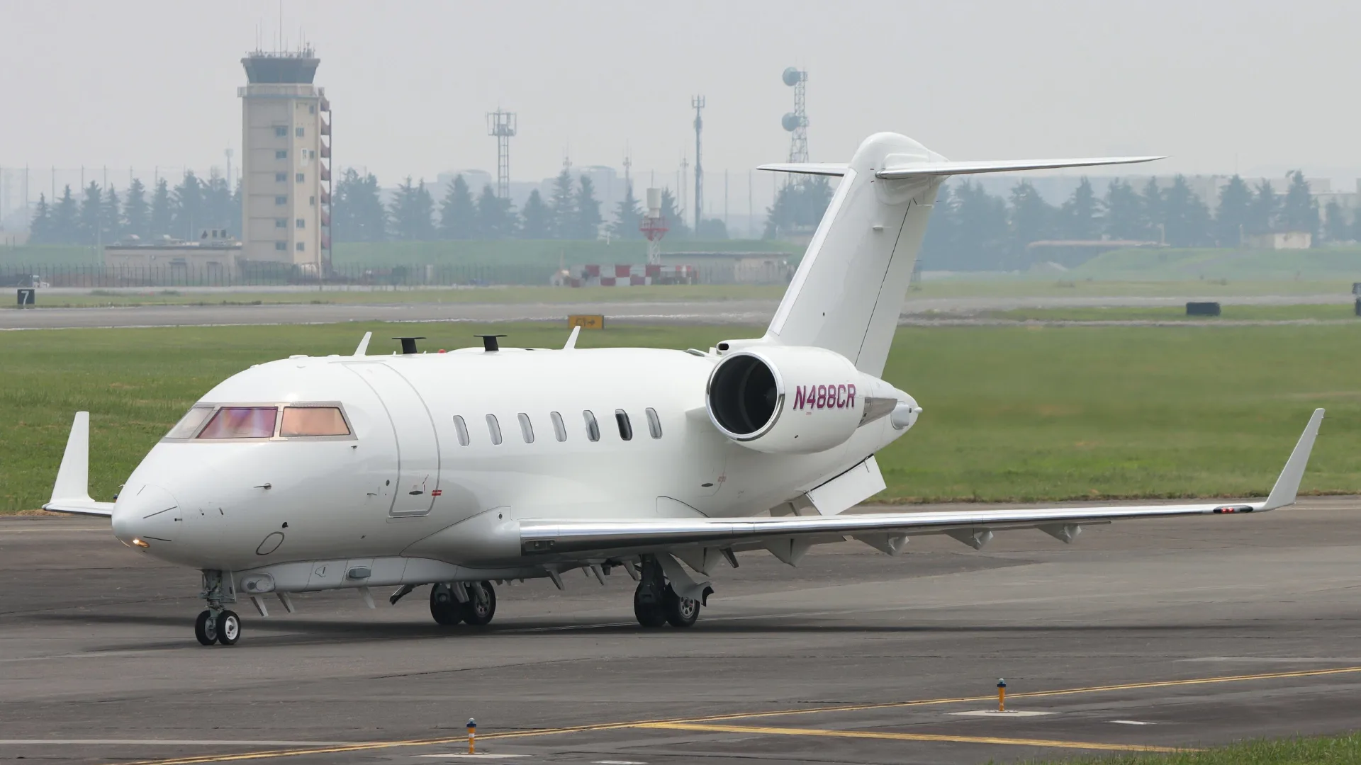 A white Bombardier Challenger 605 business jet, registration N488CR, is seen on an airport tarmac with a control tower in the background.