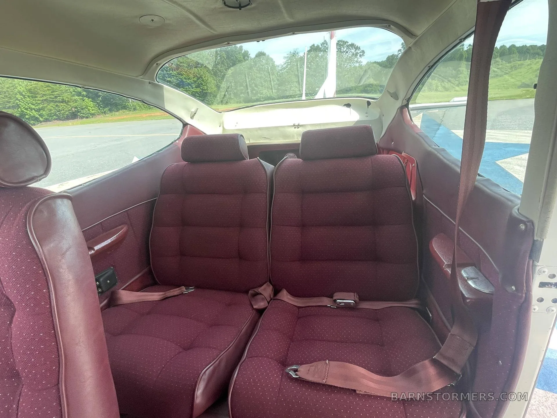 The rear cabin of the aircraft shows two maroon fabric seats with seatbelts and windows looking out onto an airfield.