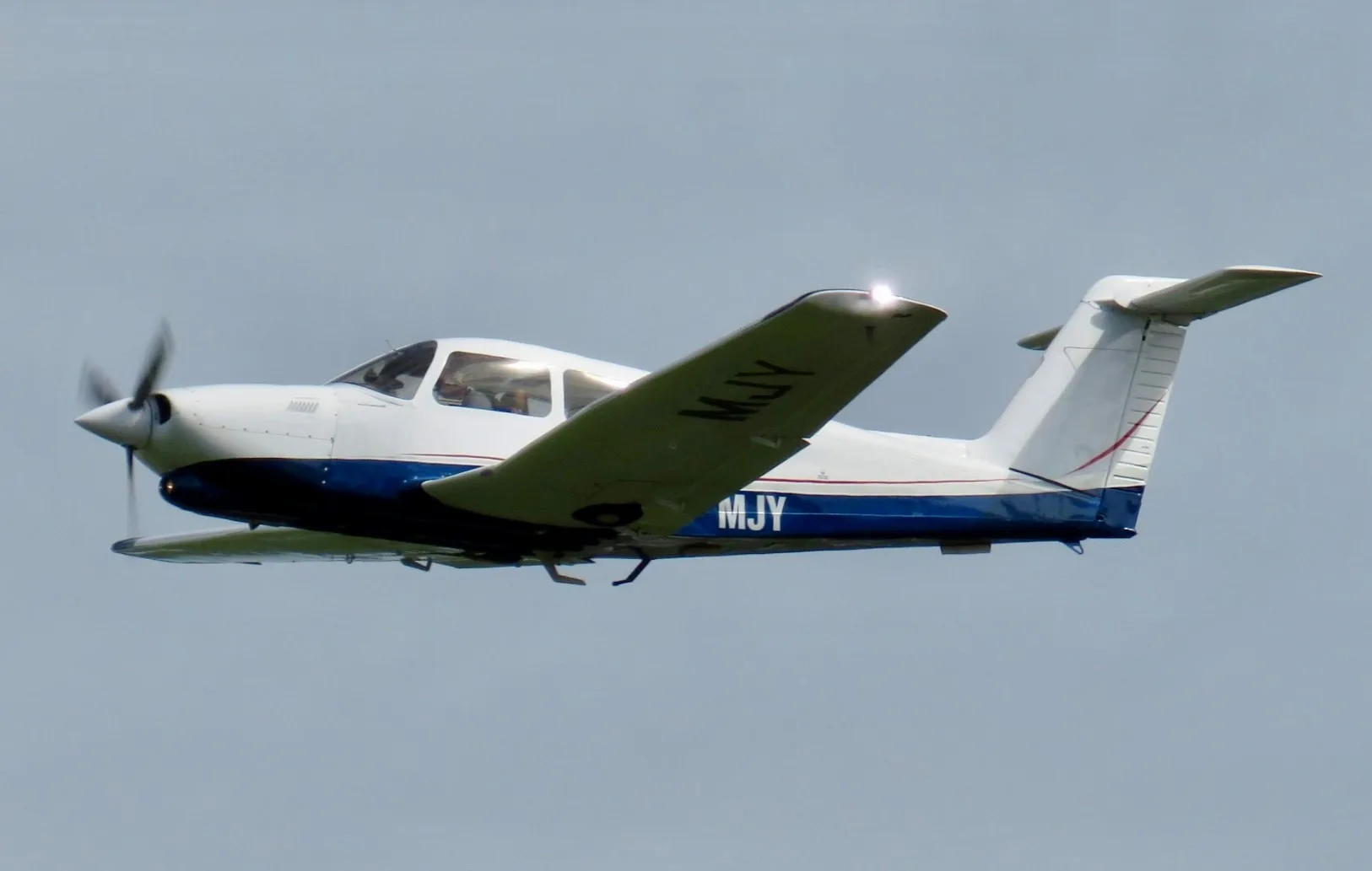 A white and blue Piper Arrow aircraft with registration MJY flying in a clear sky.