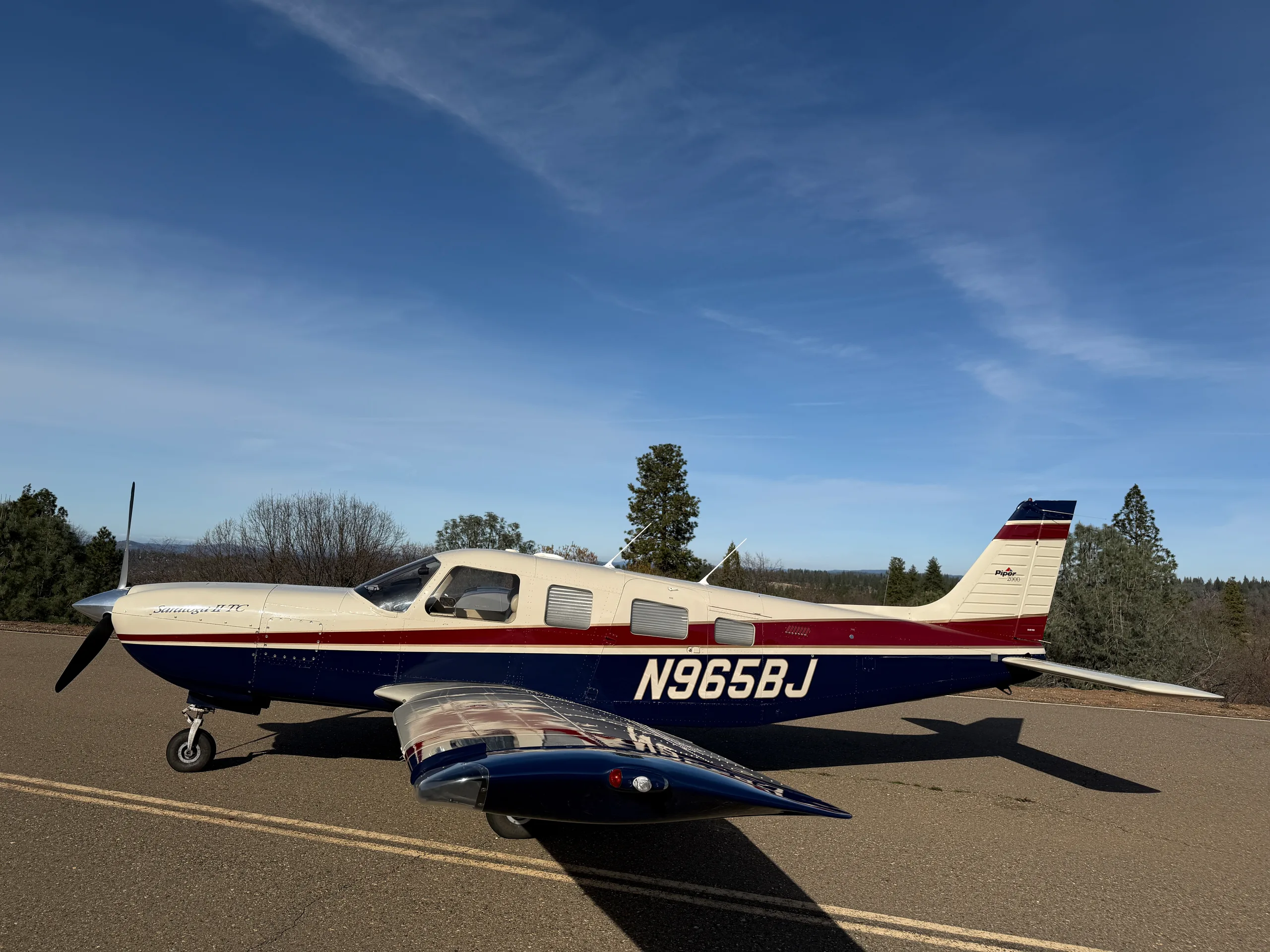 A cream, dark blue, and red Piper Saratoga II TC aircraft is parked on an asphalt surface under a clear blue sky.